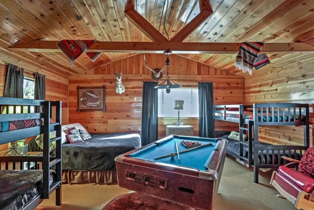 The image showcases a warm, rustic interior with a wooden ceiling, a pool table as the playful centerpiece, and cozy bunk beds along the wall. Decorative touches add charm, creating an inviting space for relaxation and family-friendly fun. The scene hints at laughter-filled pool games, comfortable sleepovers, and a welcoming atmosphere.