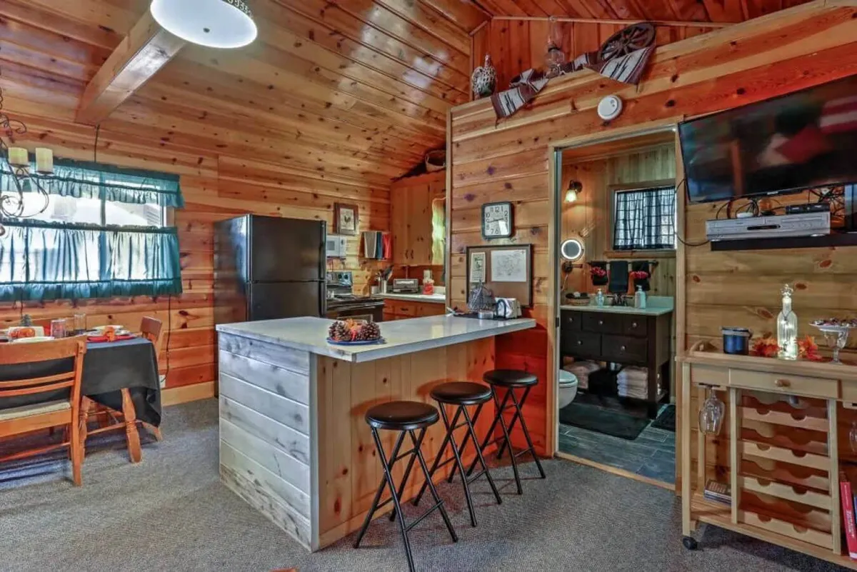 A bright, welcoming open-plan living space featuring a kitchen island, dining table, and cozy living area. The design suggests communal moments and easy transitions between indoor and outdoor activities, perfect for families and groups with pets. Large windows hint at outdoor access and natural light.
