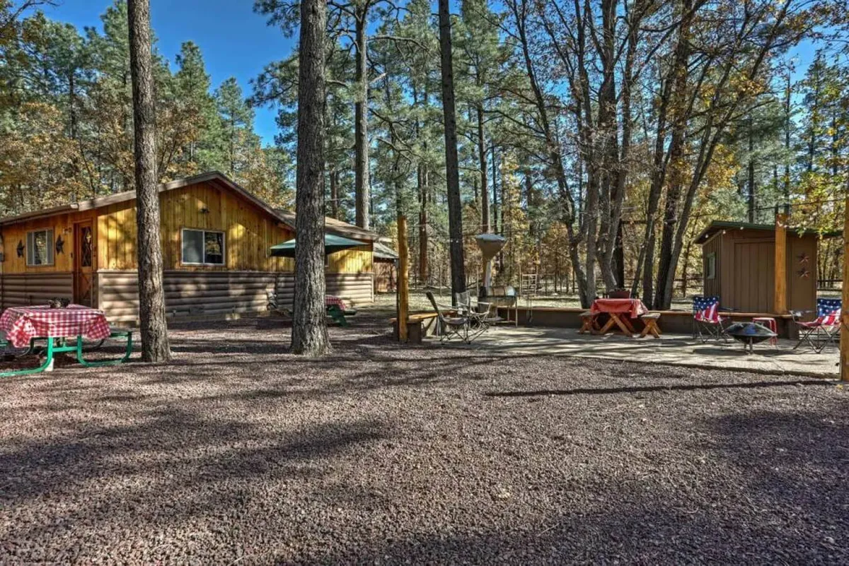 An inviting outdoor scene with a wooden cabin, picnic table, and a fire pit surrounded by trees. Sunset light glows over the fire pit area, suggesting opportunities for outdoor dining, marshmallow roasting, or stargazing after a feast.
