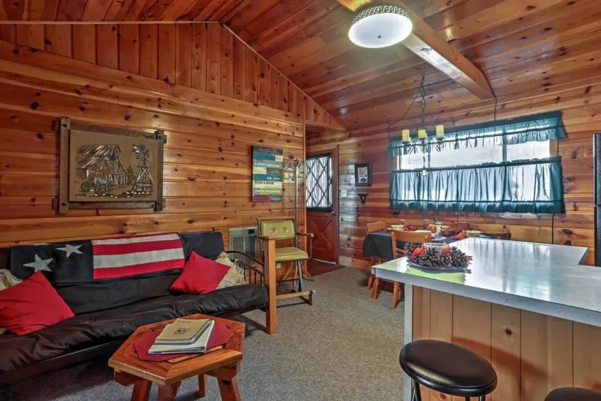 A welcoming interior space with natural wood walls, a comfortable sofa with plump cushions, a kitchen bar area, and decorative art. The setting invites socializing with friends and family, relaxing with a book, or entertaining guests in a cozy environment.