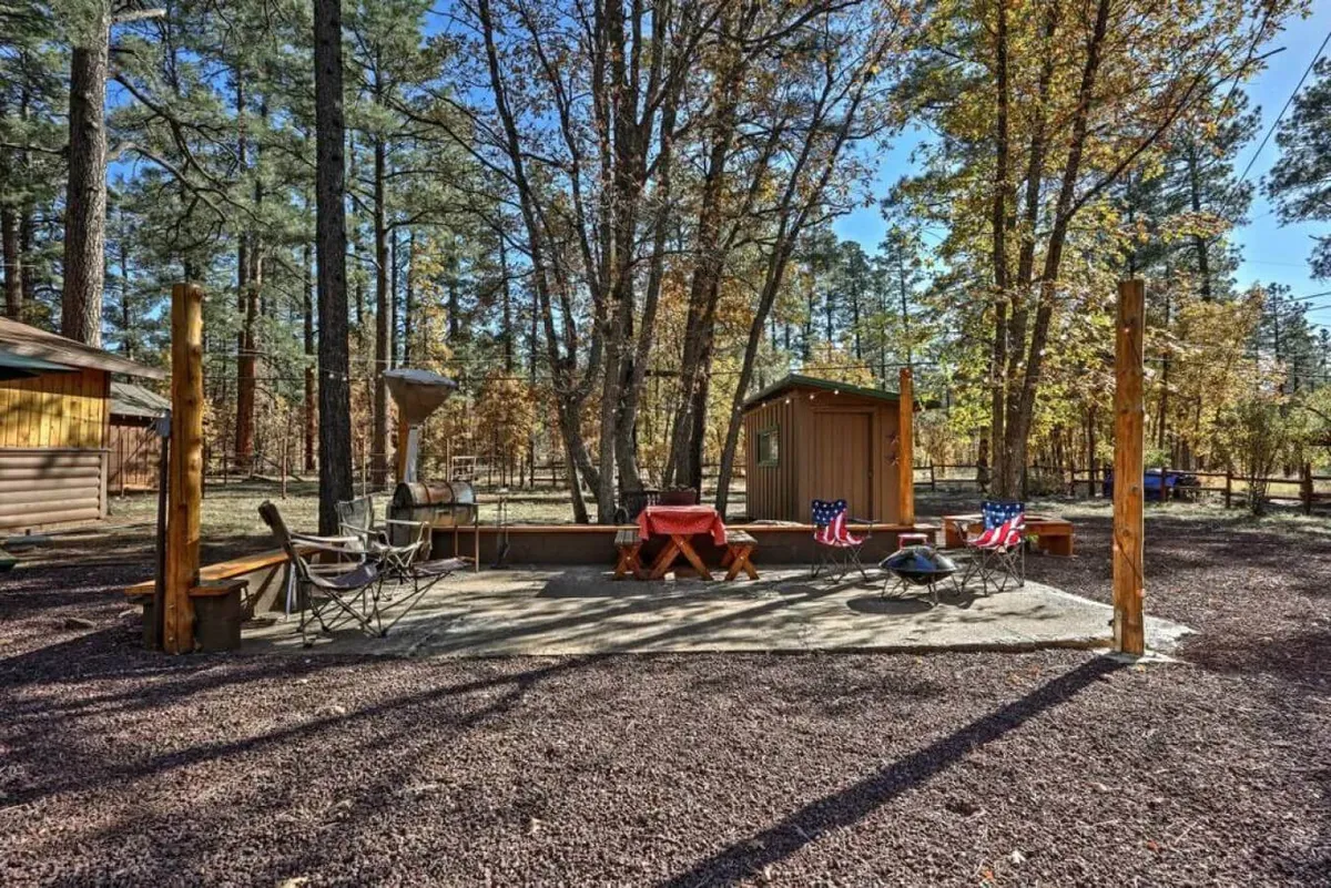 A wooden deck features comfortable outdoor chairs and a table set with a red tablecloth, overlooking a small cabin and surrounded by greenery. The area is perfect for al fresco dining, evening gatherings, or simply taking in the fresh air. The casual setup hints at shared meals and celebrations outdoors.