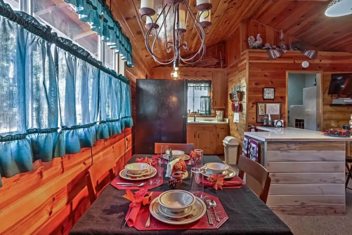 This interior image features a dining table set with plates and decor, a chandelier overhead, and blue curtains framing the window. The warm lighting and inviting arrangement create a communal atmosphere ideal for group gatherings.