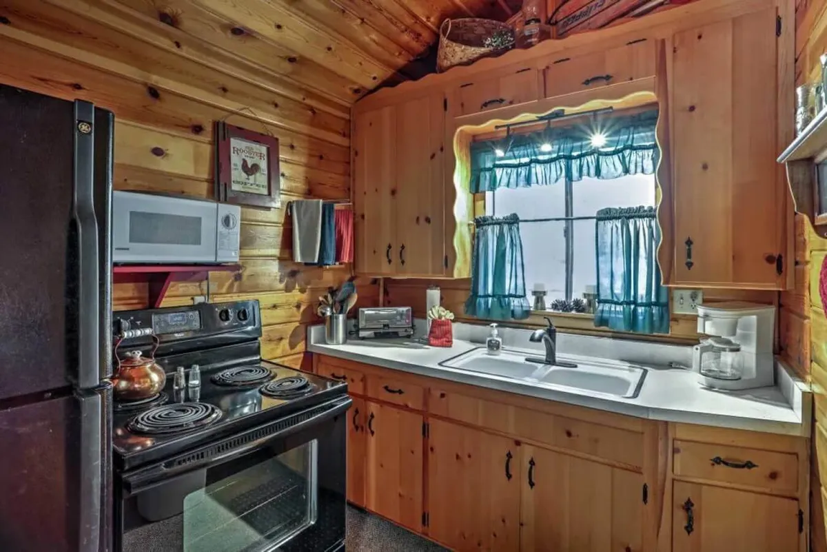 A warm and inviting kitchen featuring wooden cabinetry, a stove and oven, a sink area, and a window with curtains that lets in natural light. The setup suggests plenty of space for meal preparation and gathering for morning coffee or family cooking sessions.