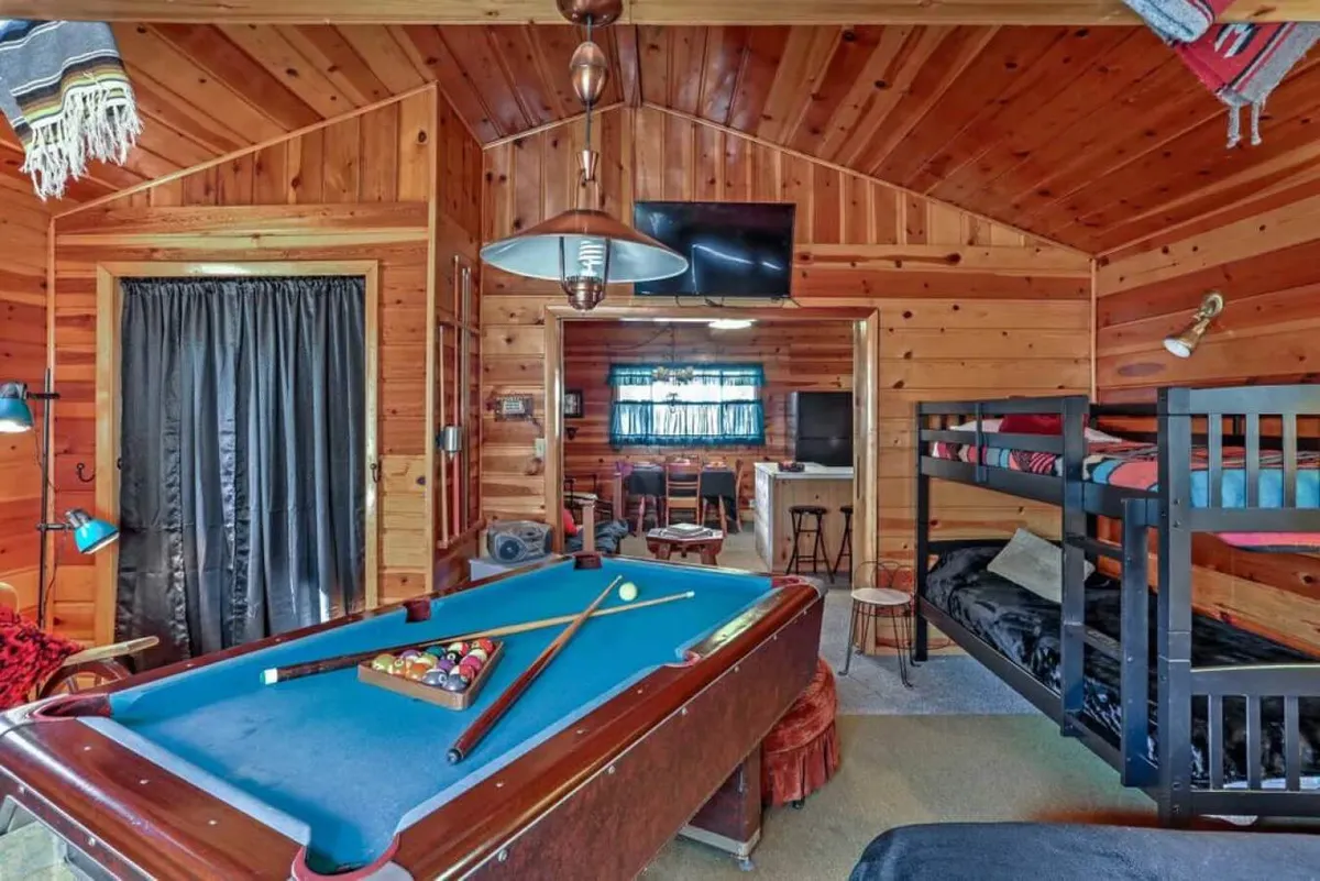 This image features a lively entertainment area with a pool table front and center, surrounded by cozy bunk beds against wood-paneled walls. The room includes a TV mounted on the wall, completing the entertainment setup. The space is well-lit and exudes a playful, communal atmosphere, perfect for group gatherings and shared relaxation.