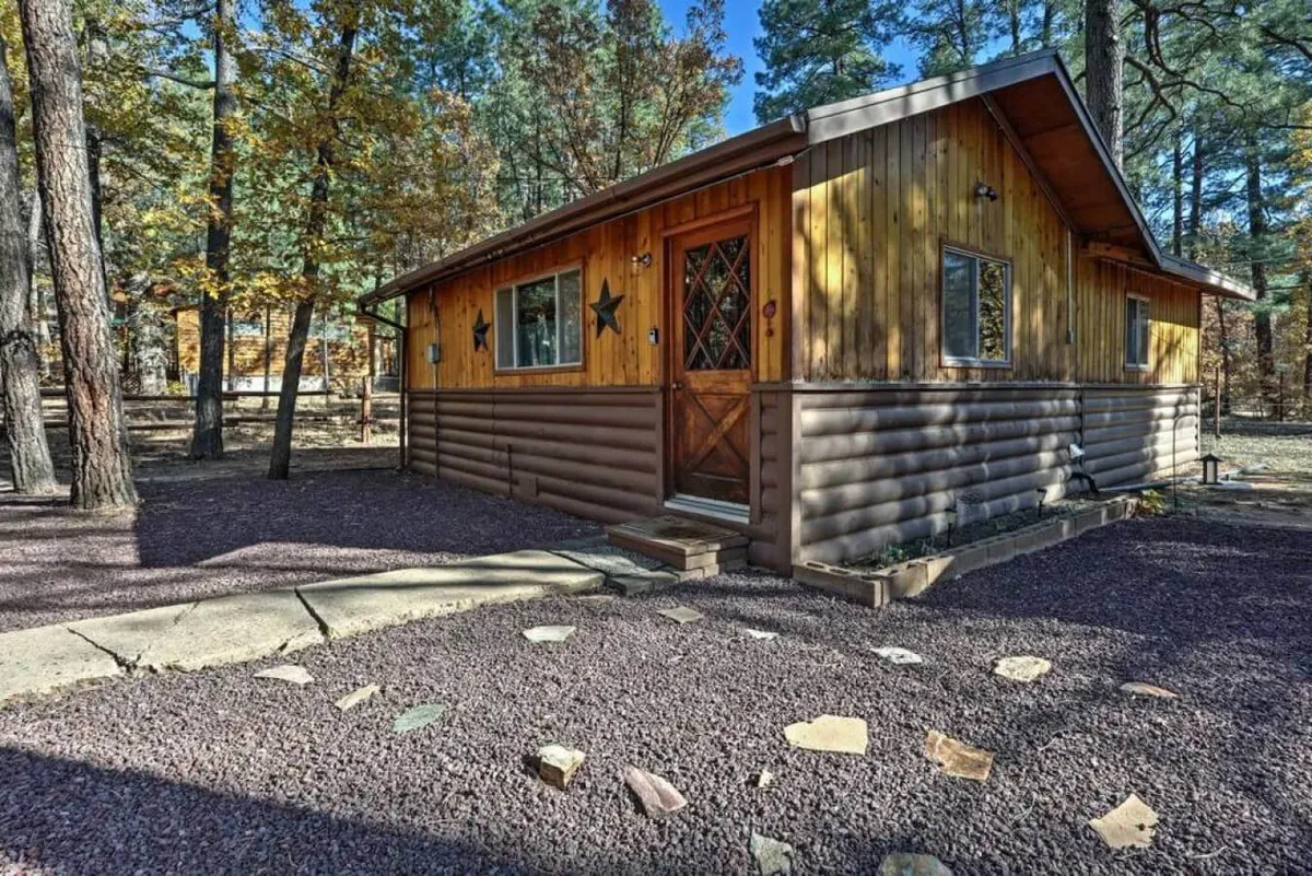 This image features the wooden exterior of the property nestled among tall trees, with a welcoming front door adorned with a diamond window and decorative stars. The surrounding greenery suggests immediate access to nature and outdoor adventure just steps away from the cabin.