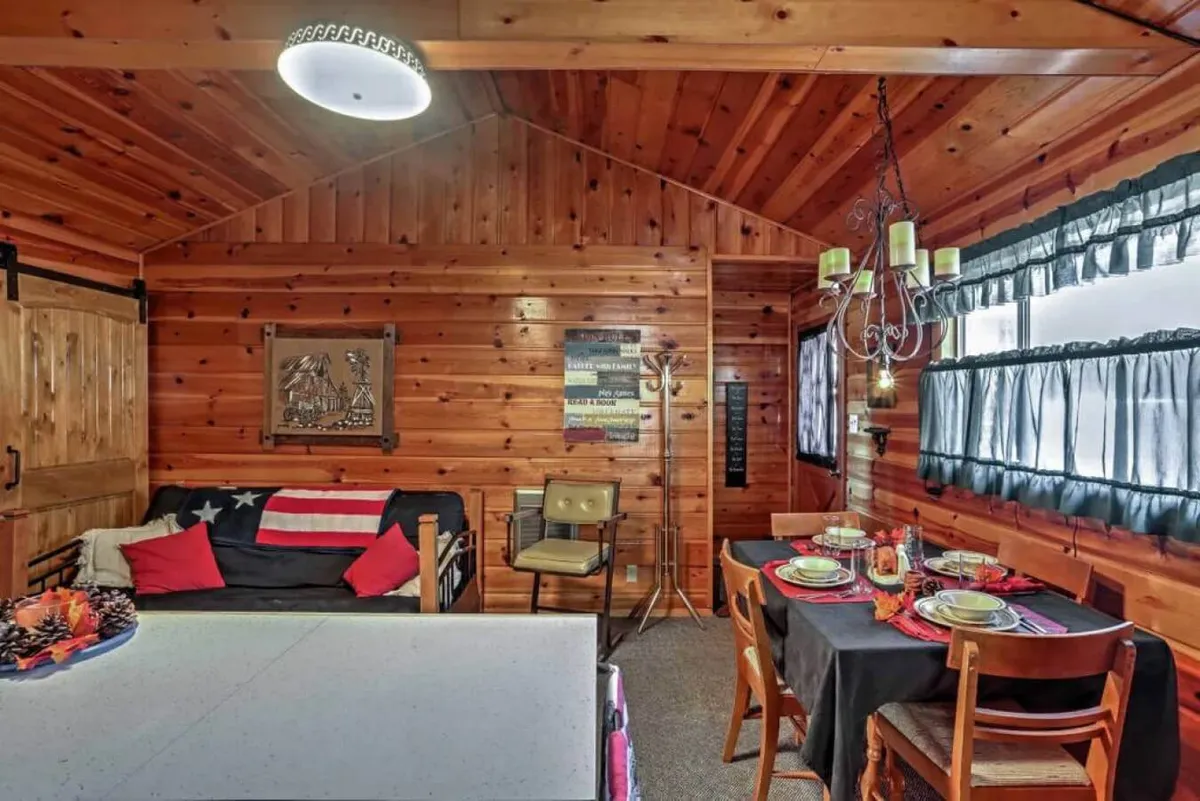 A warm, wood-paneled common area featuring an inviting seating space adorned with American flag decor. A dining table is set for a meal, with a chandelier overhead and decorative wall art in the background. The scene evokes shared laughter, family meals, and casual gatherings, creating a camp-like, communal atmosphere.